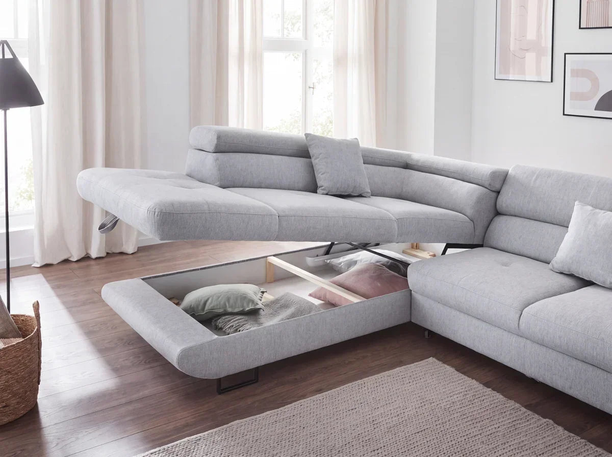A light gray fabric sectional sofa shown in a bright living room, with the front cushion pulled down to reveal storage space underneath. The room has wood floors, beige curtains, and a woven rug.